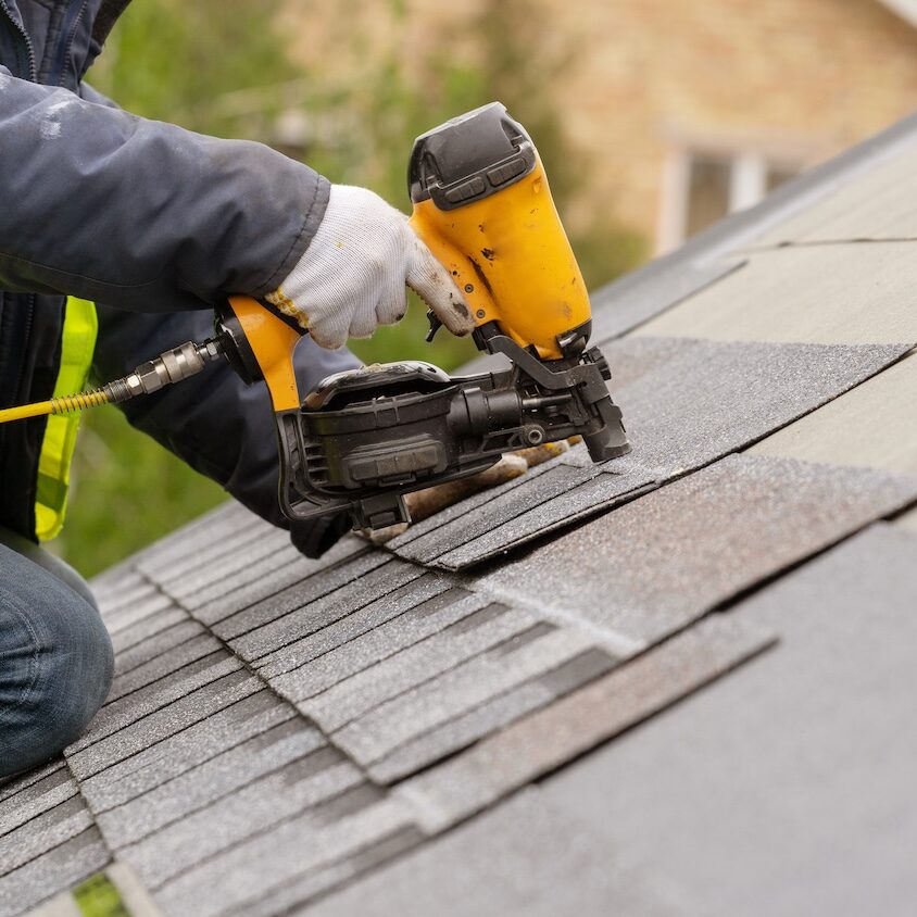Roofer worker in special protective work wear and gloves, using air or pneumatic nail gun and installing asphalt or bitumen shingle on top of the new roof under construction residential building