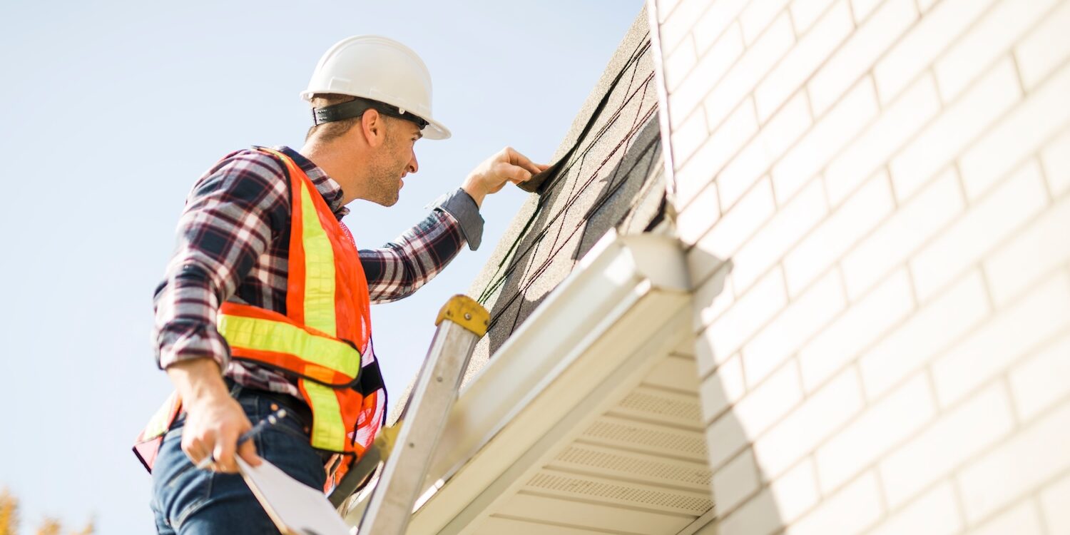 A man with hard hat standing on steps inspecting house roof