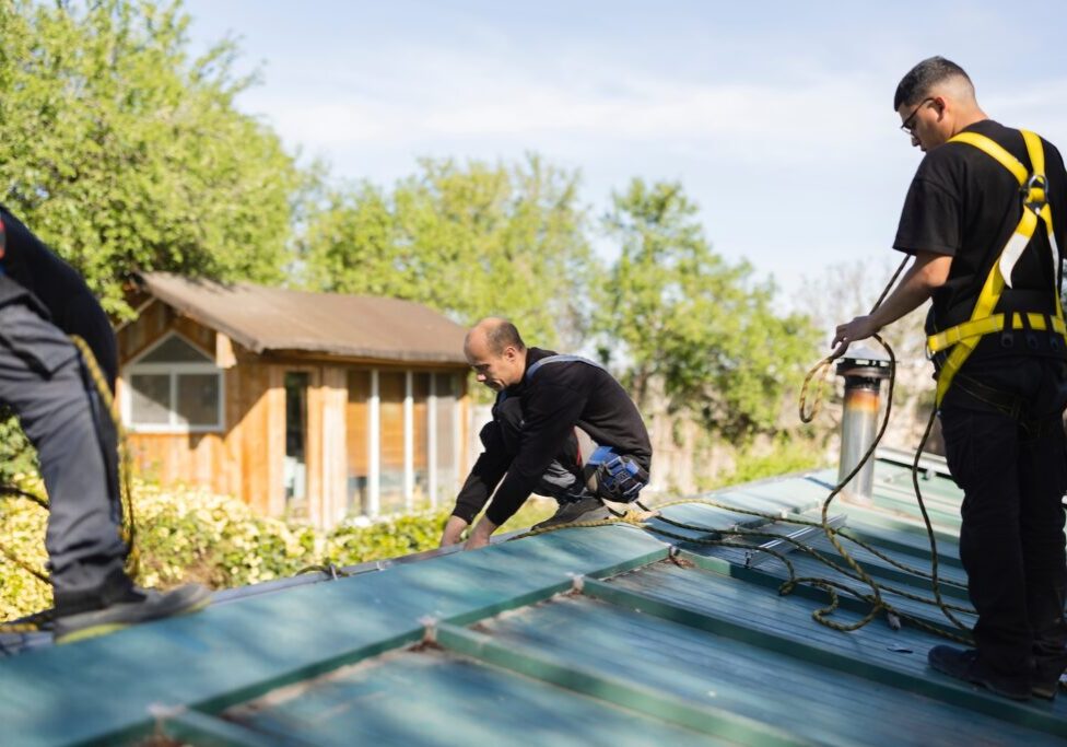 Construction workers securing themselves with safety harnesses while installing metal roof sheets on a house, ensuring workplace safety during roofing work