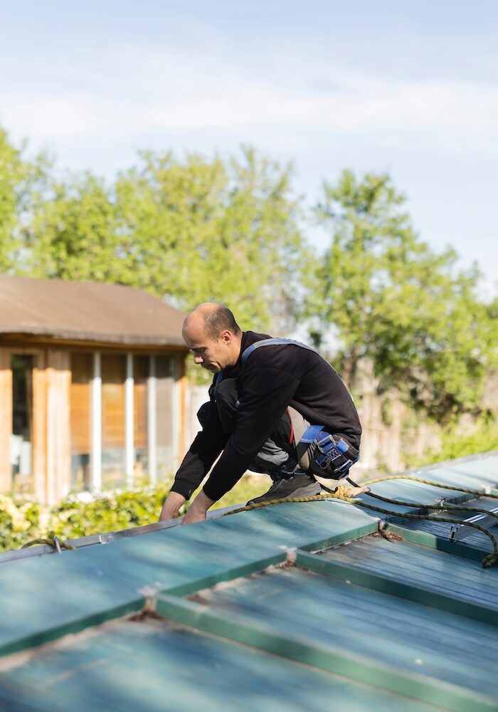 Construction workers securing themselves with safety harnesses while installing metal roof sheets on a house, ensuring workplace safety during roofing work