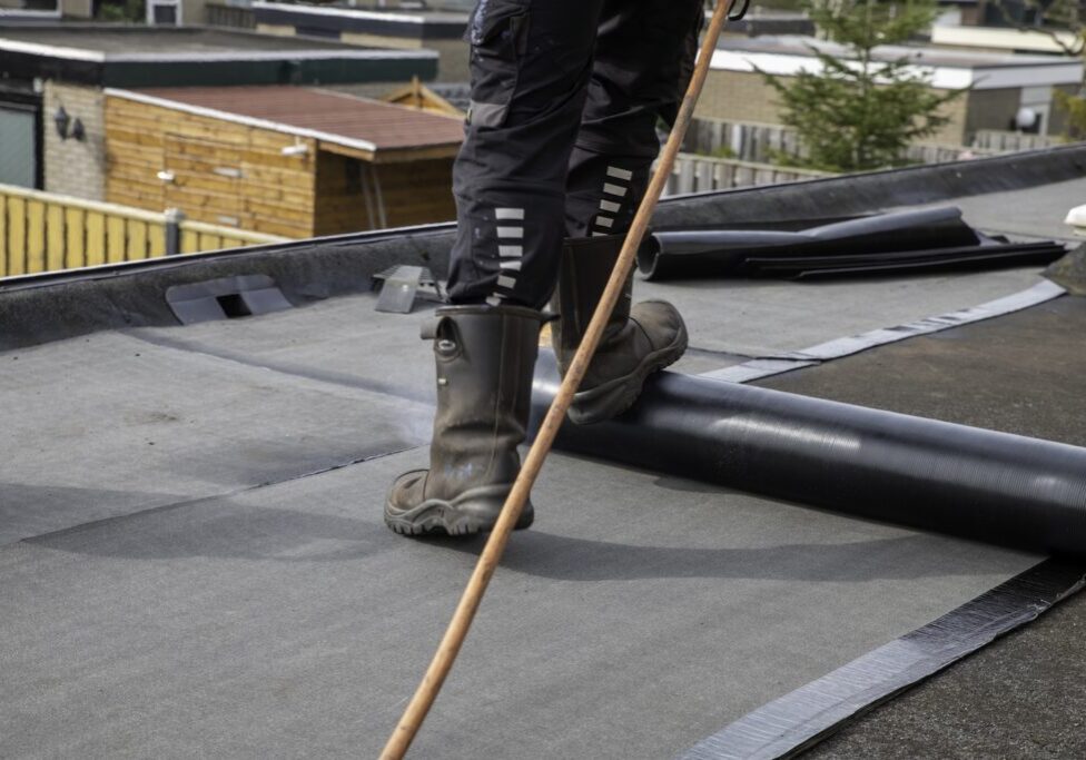 Roofer standing on a flat residential roof using a propane torch to heat and install roll roofing membrane for waterproofing and roof repair. Scene shows work boots, tools and neighborhood houses.