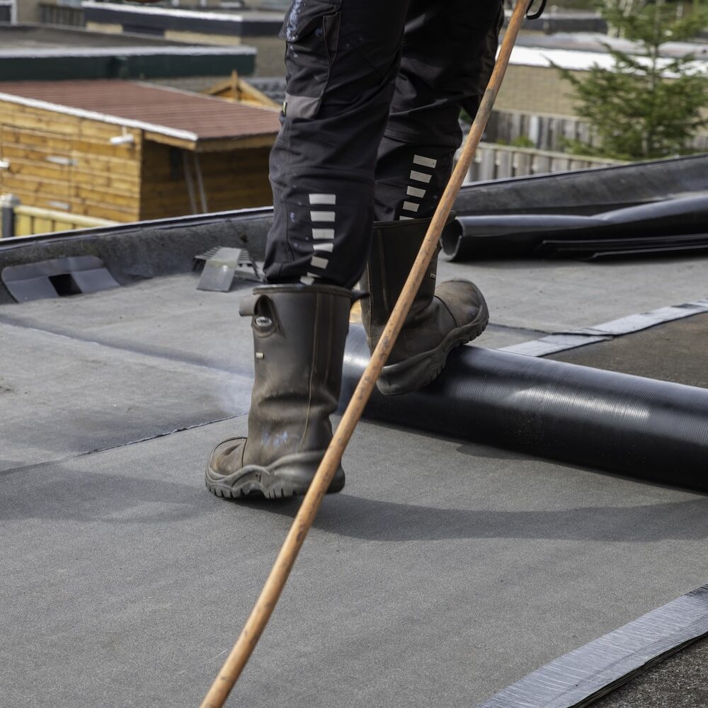 Roofer standing on a flat residential roof using a propane torch to heat and install roll roofing membrane for waterproofing and roof repair. Scene shows work boots, tools and neighborhood houses.